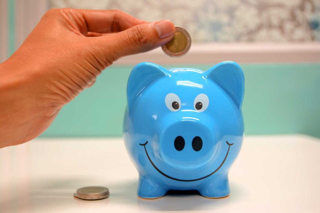 A Hand Placing A Coin In A Blue Piggy Bank.