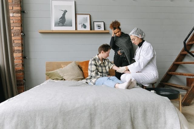 Nurse Talking To A Woman Lying In A Bed While Being Comforted By Her Friend As An Example Of Liposculpture Post-Operative Care