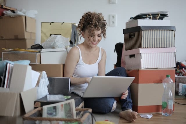 Woman Sitting On The Floor Between Boxes Of Stuff And Looking At Her Laptop